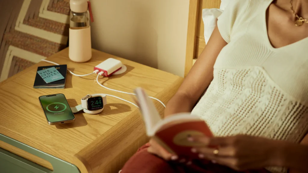 Ein iPhone und eine Apple Watch laden gleichzeitig über das kompakte „ButterFly“-Ladegerät von Twelve South auf einem Nachttisch, während eine Person im Bett ein Buch liest.