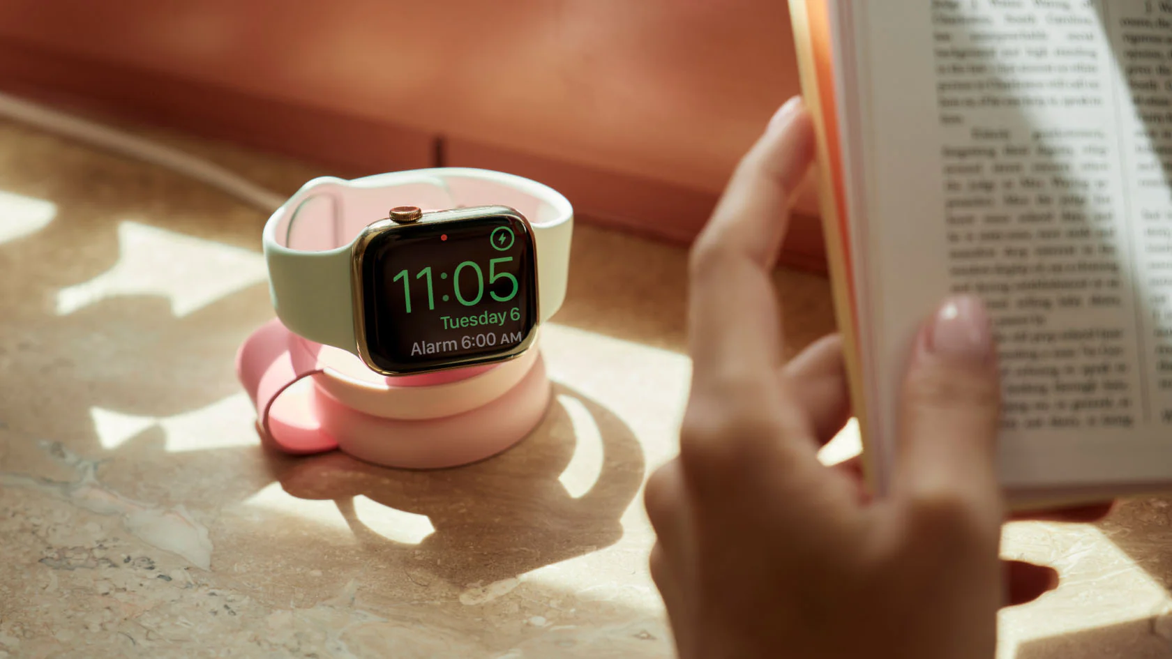 Eine Apple Watch lädt auf dem rosa „ButterFly“-Ladegerät von Twelve South, während im Vordergrund eine Person ein Buch liest.