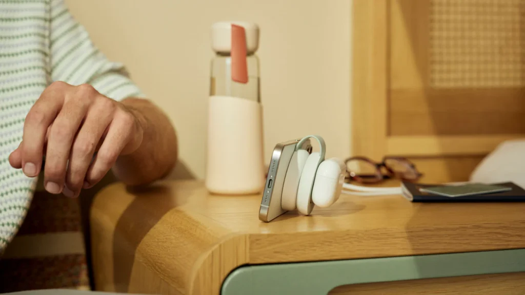 Ein Smartphone lädt im Standmodus auf dem kompakten „ButterFly“-Ladegerät von Twelve South, während daneben kabellose Ohrhörer auf einem Holztisch liegen.
