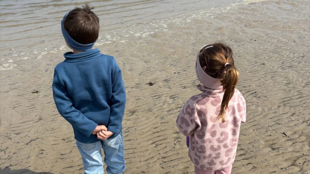 Zwei Kinder in Gummistiefeln stehen am Nordseestrand und blicken auf das ruhige Wattenmeer bei Ebbe.