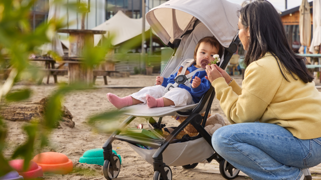 Ein Kleinkind sitzt entspannt im Joolz Aer² Buggy in der Farbe Calming Beige auf einem Spielplatz, während eine Person ihm eine Blume zeigt.