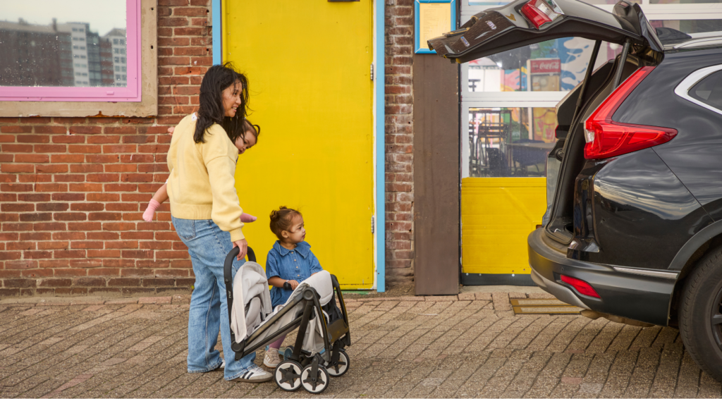 Eine Person steht mit zwei Kindern neben einem Auto und einem zusammengefalteten Joolz Aer² Buggy in der Farbe Calming Beige vor einem Gebäude mit gelber Tür.