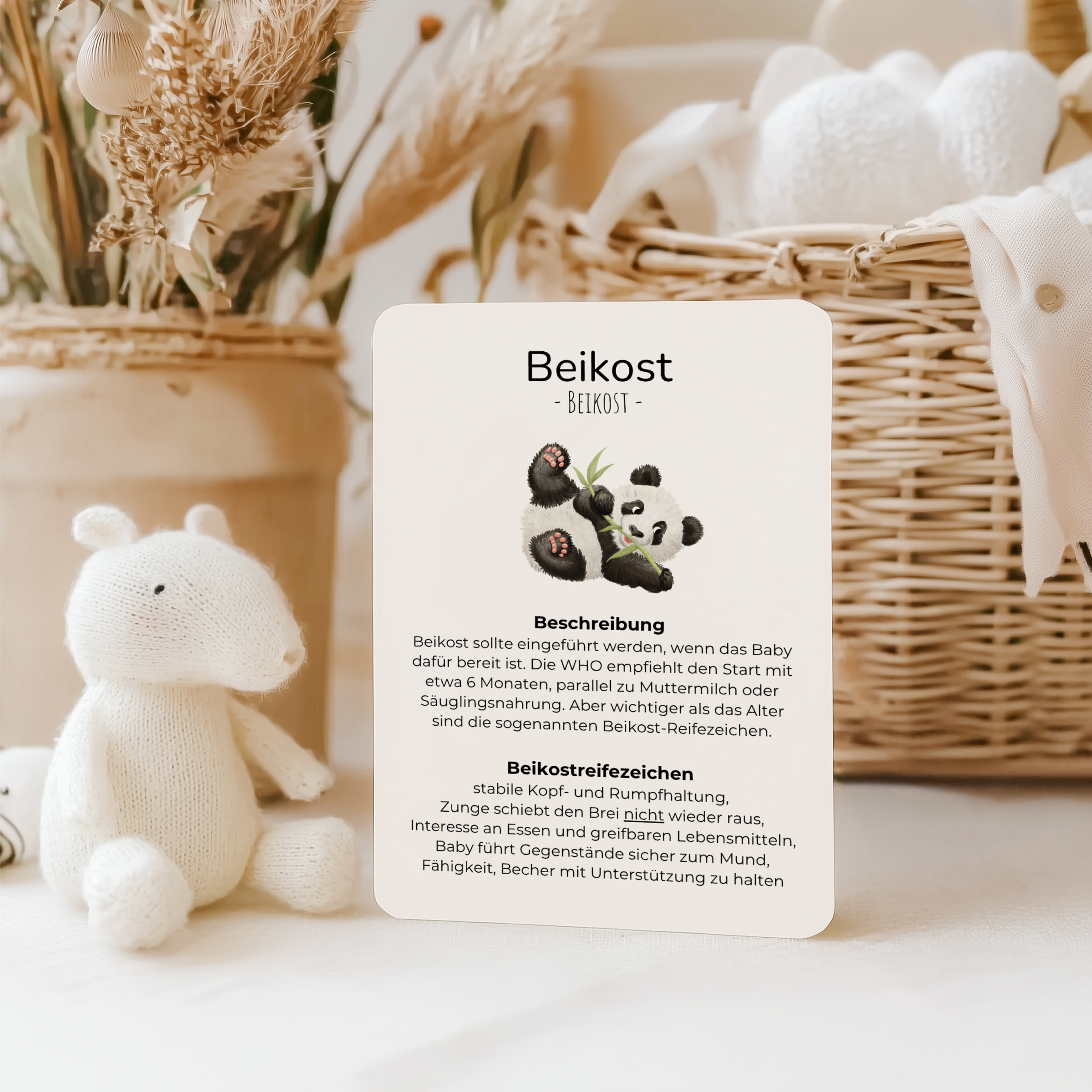 Eine Informationskarte zum Thema Beikost aus einem Elternratgeber-Kartenset steht auf einem hellen Tisch in einer liebevoll gestalteten Babyumgebung mit Kuscheltier, Korb und natürlichen Dekoelementen. Die Karte informiert über den Beikoststart und typische Beikostreifezeichen.