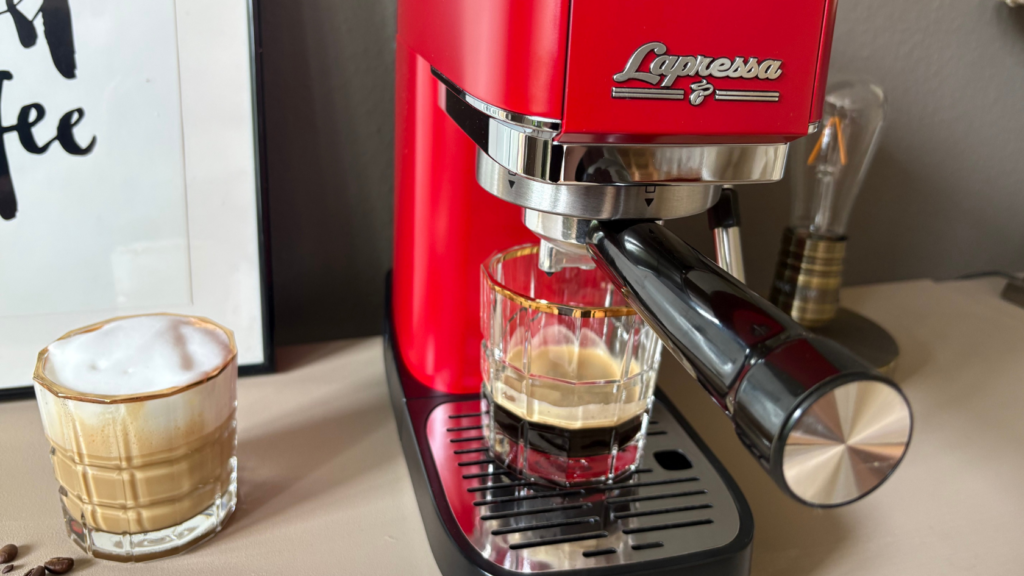 Nahaufnahme der roten Siebträgermaschine Tchibo Lapressa während der Espressozubereitung. Unter dem Siebträger steht ein Glas mit frisch gebrühtem Espresso und heller Crema. Daneben befindet sich ein cremiger Cappuccino mit feinporigem Milchschaum auf einer hellen Arbeitsfläche.