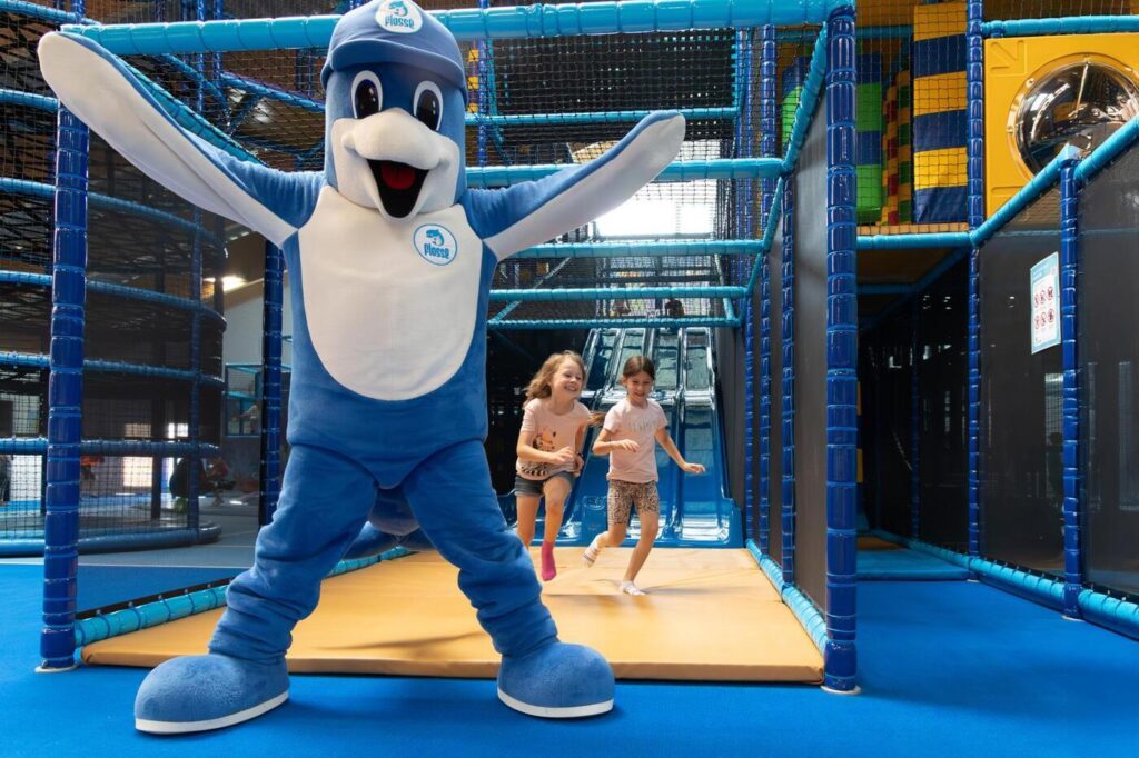 Zwei Kinder spielen gemeinsam mit dem Maskottchen Flosse im großen Indoor-Spielpark des Flosse Abenteuerlands im Aldiana Club Ampflwang.