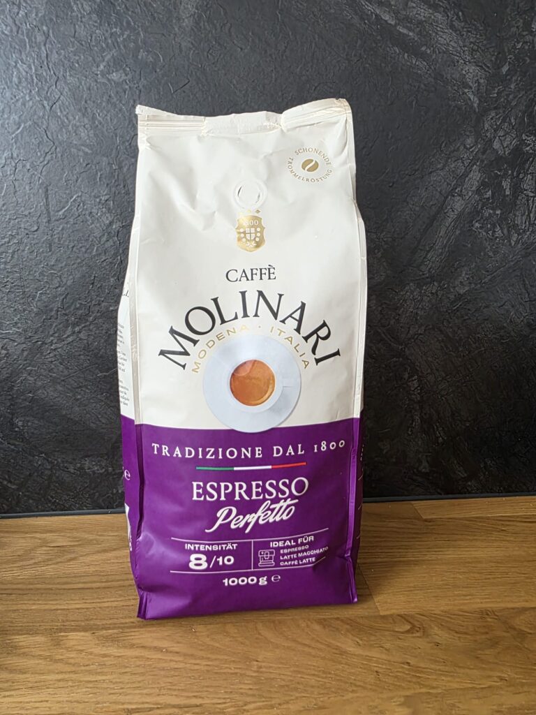Eine Packung Caffè Molinari Espresso Perfetto mit ganzen Kaffeebohnen steht auf einer Holzarbeitsplatte vor einem dunklen Hintergrund. Die Verpackung ist weiß-violett gestaltet und trägt den Hinweis „Tradizione dal 1800“. Der Kaffee eignet sich laut Verpackung besonders für Espresso, Latte Macchiato und Caffè Latte.