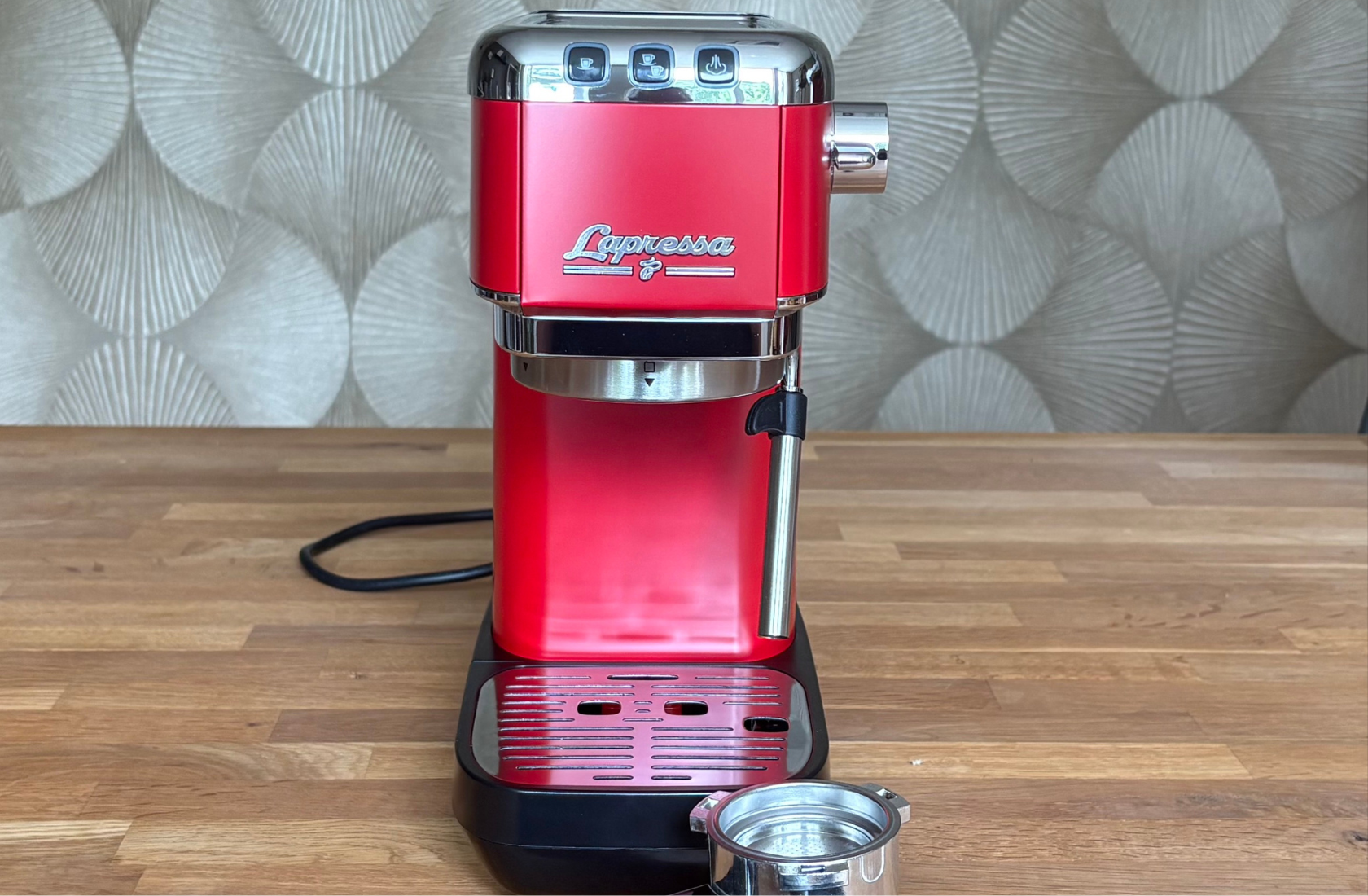 Die rote Siebträgermaschine Tchibo Lapressa steht auf einer Holzarbeitsplatte vor einer dezent gemusterten Wand. Vor der Maschine liegt der Edelstahl-Siebträger mit Filtersieb, wodurch das kompakte Design und die Ausstattung der Espressomaschine gut zu erkennen sind.
