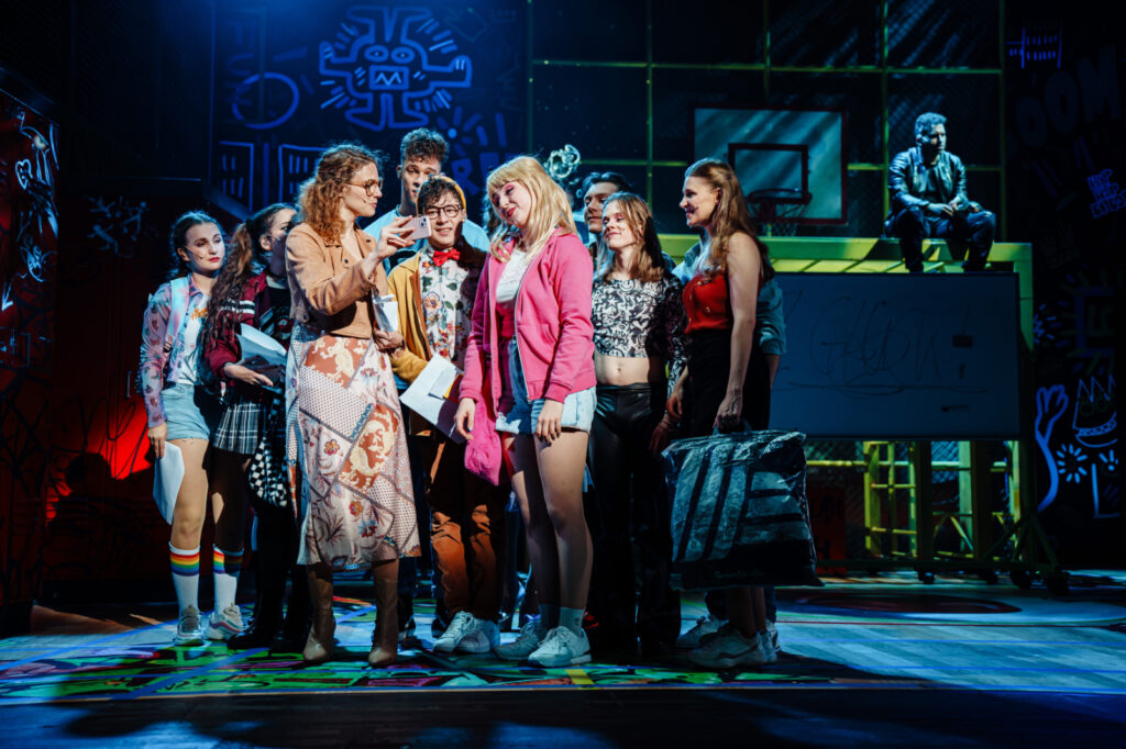 Szene aus FACK JU GÖHTE – Das Musical: Mehrere Darstellerinnen und Darsteller stehen als Schulklasse auf einer modernen Bühne mit Graffiti-Kulisse. Im Mittelpunkt zeigt eine Lehrerin den Jugendlichen etwas auf ihrem Smartphone, während im Hintergrund Zeki Müller auf einem Klettergerüst sitzt. Die lebendige Inszenierung vermittelt den typischen Mix aus Humor, Chaos und Schulalltag.