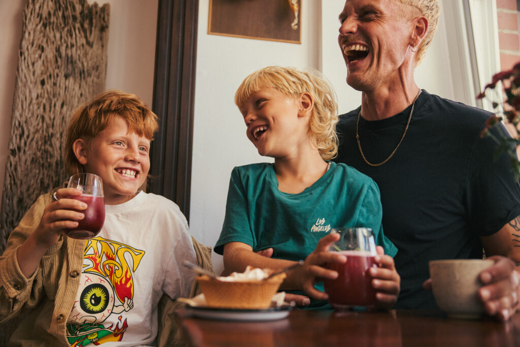 Ein Mann und zwei Kinder lachen gemeinsam an einem Tisch und halten Gläser mit rotem Getränk.
